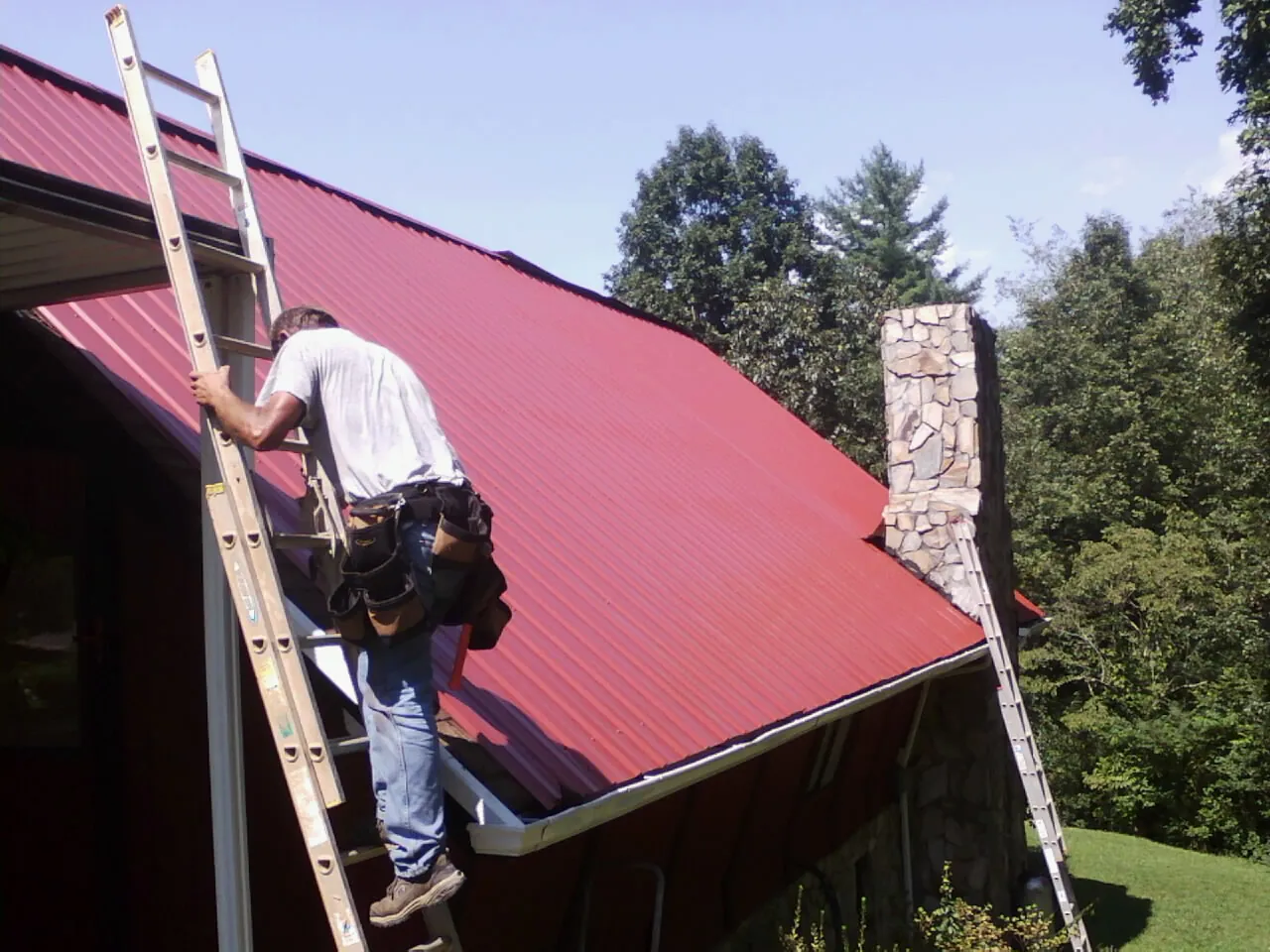 Roofer on a ladder beside a red metal roof and stone chimney — Soffit & Fascia Repair by Roofing Brothers in Rhinebeck